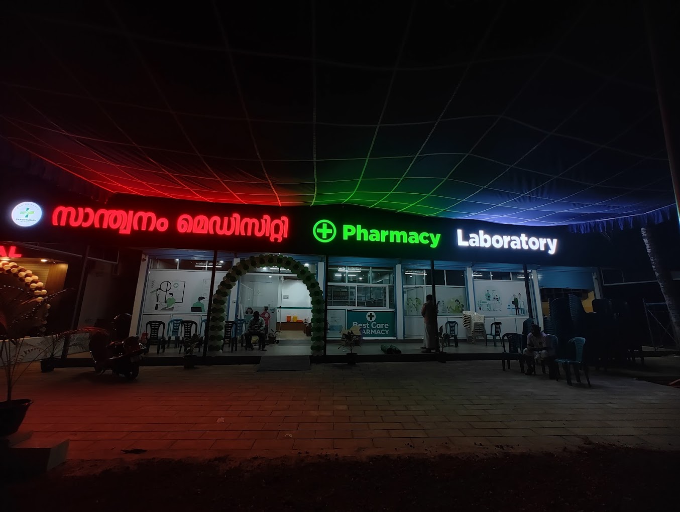 Santhwanam Medicity storefront at night, its green Pharmacy and Laboratory signs and Malayalam സാന്ത്വനം മെഡിസിറ്റി wordmark lit up.