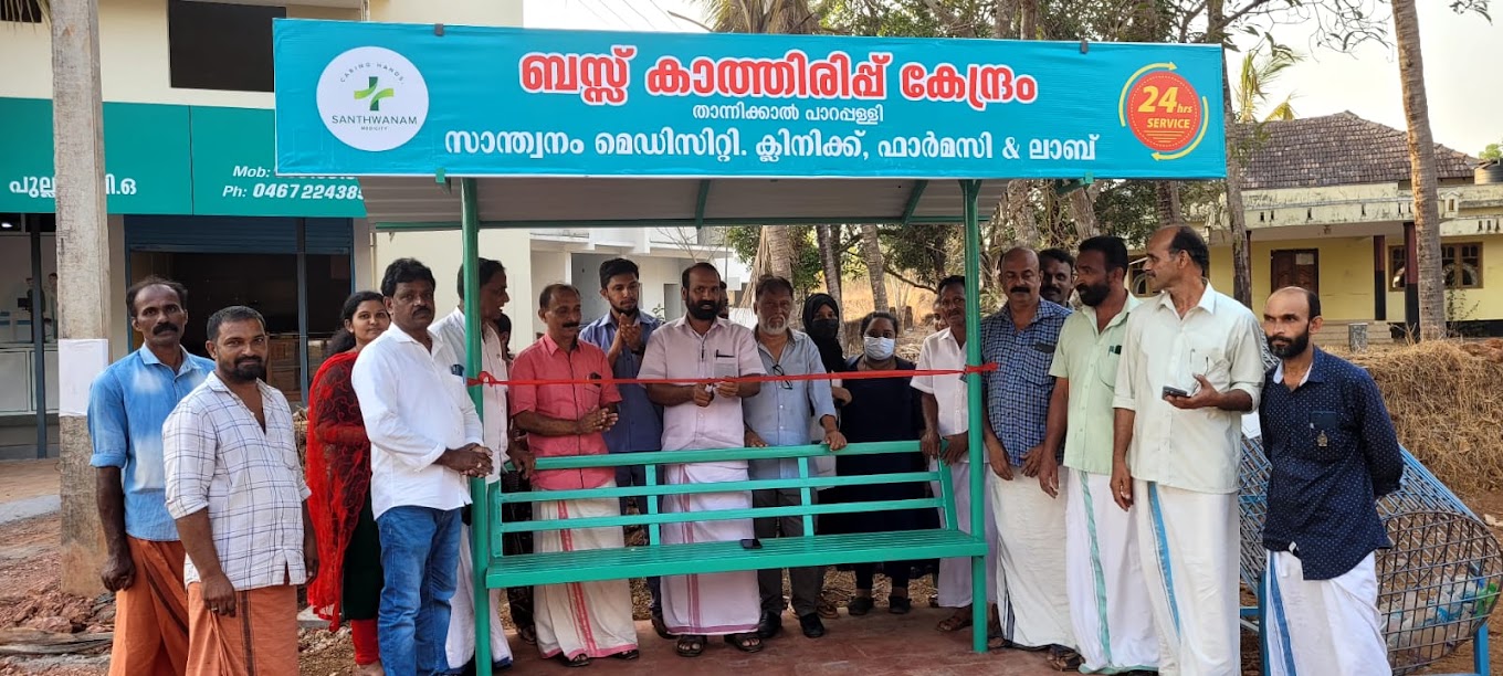 A green roadside bus-stop shelter sponsored by Santhwanam Medicity, being opened with a ribbon-cutting as neighbours gather.