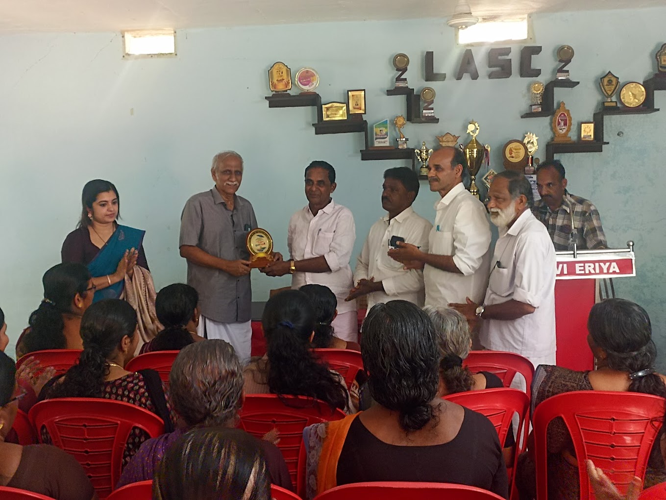 A community gathering where a memento is presented on stage, with neighbours seated together in the audience.